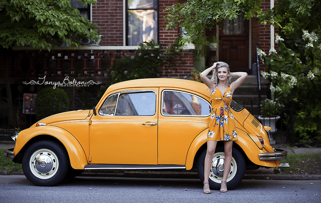 A teenager in a yellow floral dress standing in front of a matching yellow Volkswagen Bug with her hands in her hair for a senior picture.