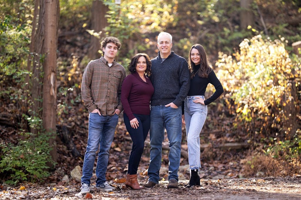 A family of four dressed in coordinating colors standing in a wooded area with colorful fallen leaves for a portrait.