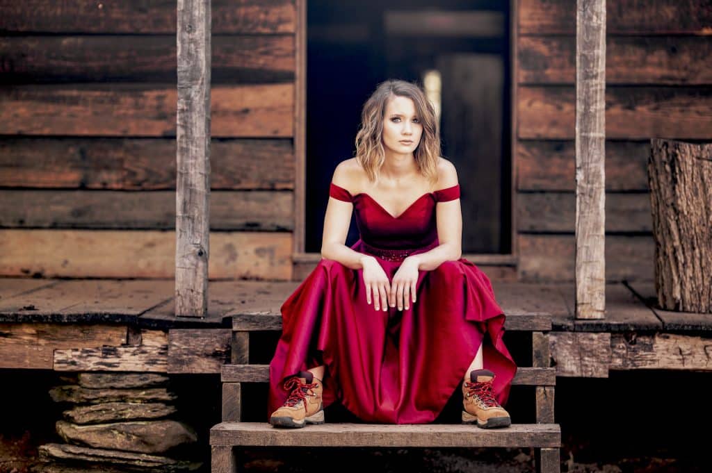 A girl in a formal red gown and old hiking boots with matching red laces sitting on the front step of a rustic cabin in Gatlinburg, Tennessee.