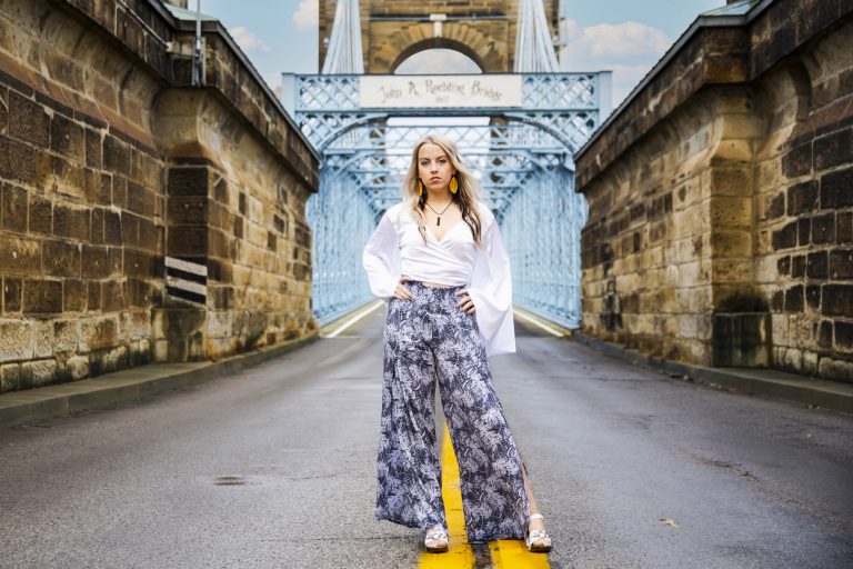 Blonde teenager standing in the middle of the street with the John R. Roebling bridge entrance behind her.