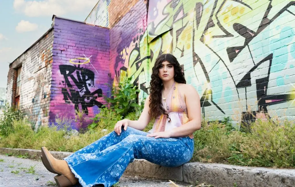 A Cincinnati senior portrait of a girl wearing flared jeans sitting on a curb in front of a building covered in colorful graffit.