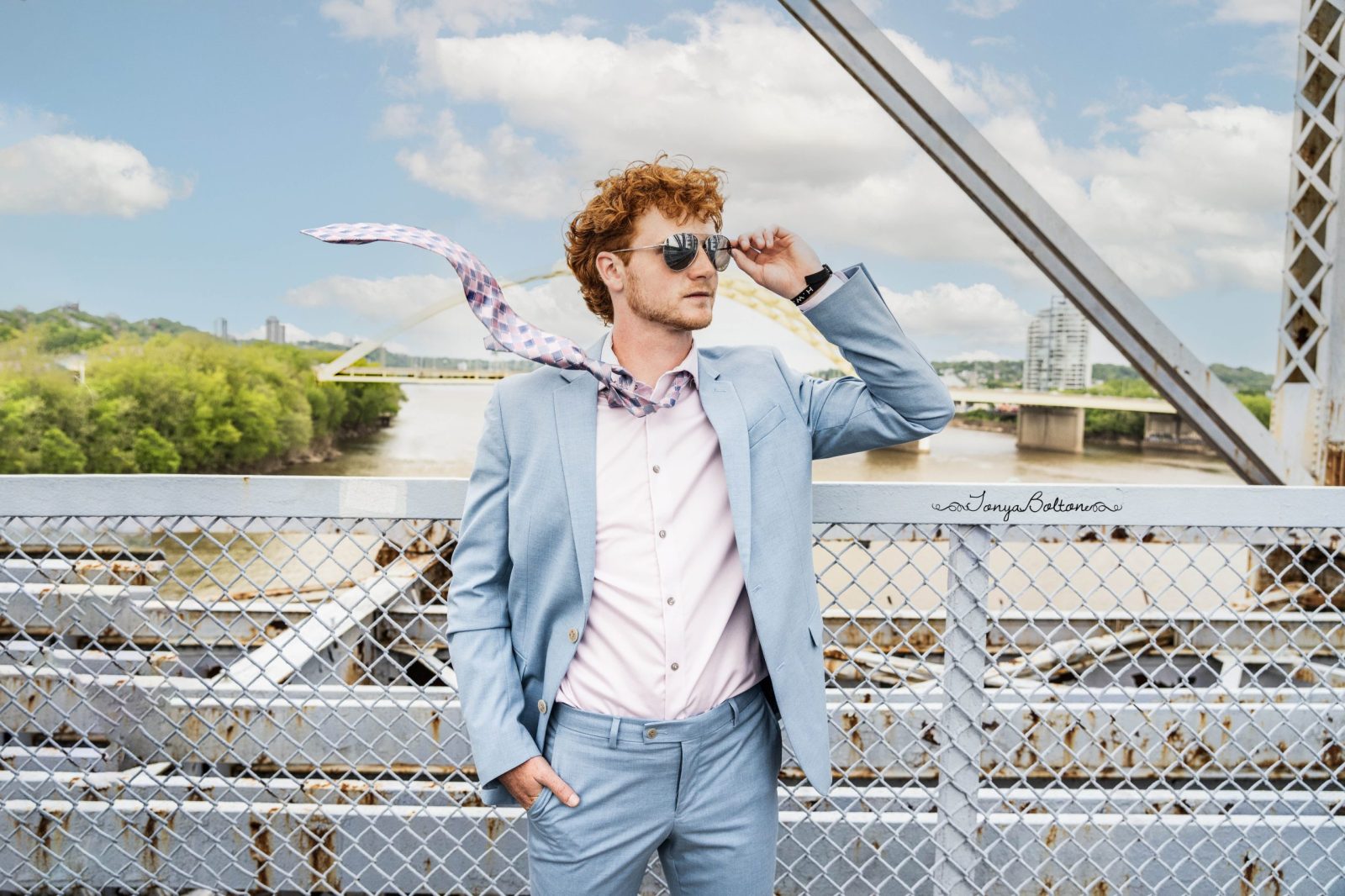 High school senior boy in a suit and sunglasses standing on the Purple People Bridge with his tie flying in the air like the wind took it while he's holding his sunglasses in Cincinnati Ohio