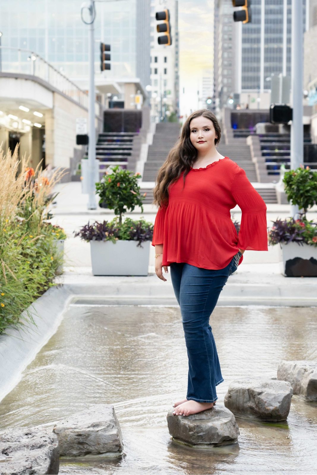 a senior portrait of a girl in a red shirt standing on a rock in front of downtown cincinnati buildings