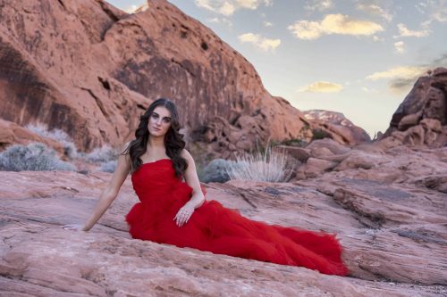 A senior portrait created by Tonya Bolton of a dark haired girl sitting in a long, red formal dress in Nevada's Valley of Fire State Park.